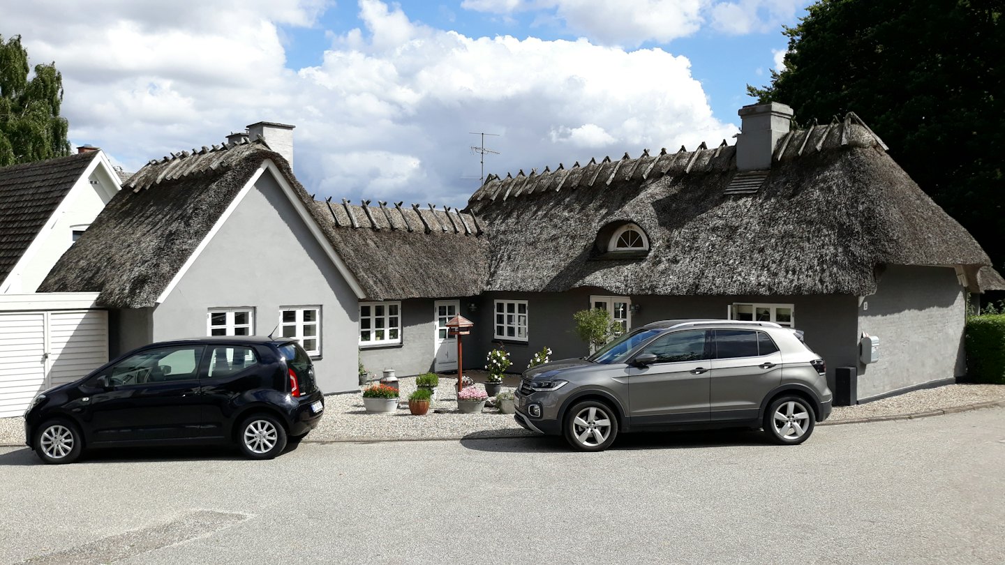 Czarny hatchback i szary SUV zaparkowane przed domem krytym strzechą.